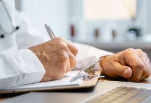 Close-up of hands writing on a clipboard with a pen, indicating professional documentation in a healthcare setting.