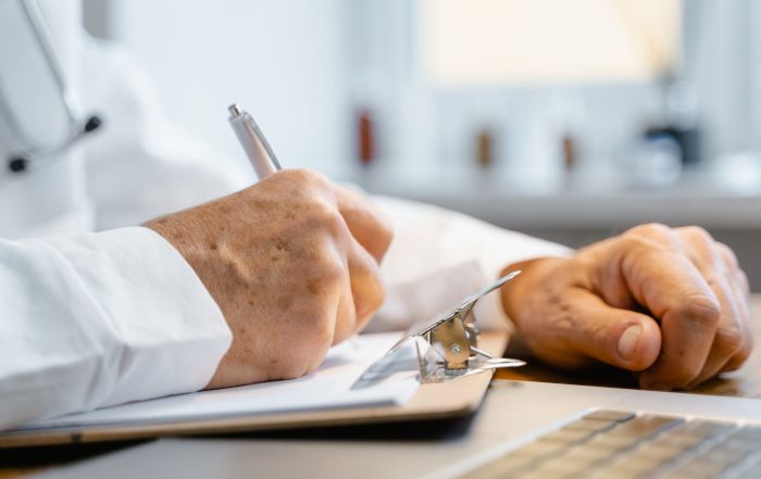 Close-up of hands writing on a clipboard with a pen, indicating professional documentation in a healthcare setting.